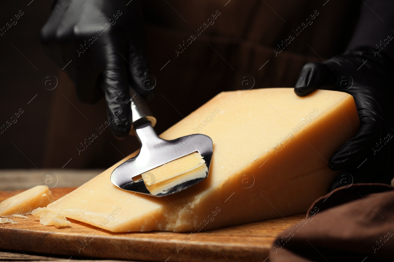 Photo of Woman in gloves cutting delicious cheese with slicer at wooden table against black background, closeup