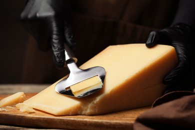 Photo of Woman in gloves cutting delicious cheese with slicer at wooden table against black background, closeup