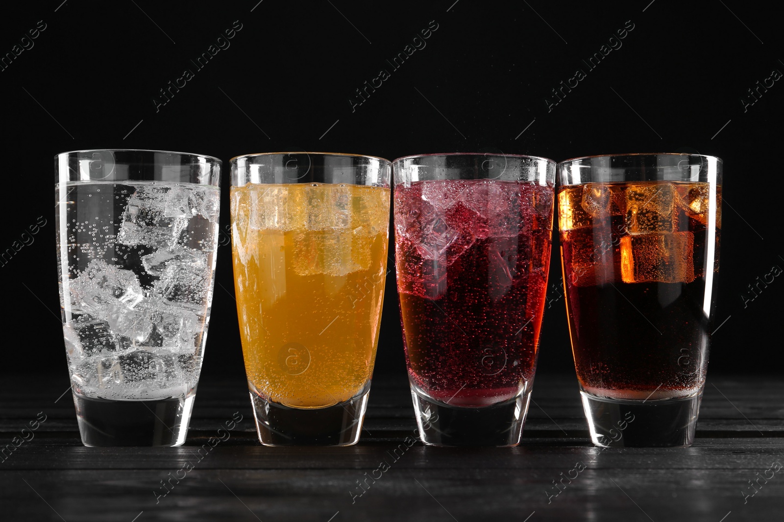 Photo of Refreshing soda water of different flavors with ice cubes in glasses on black wooden table