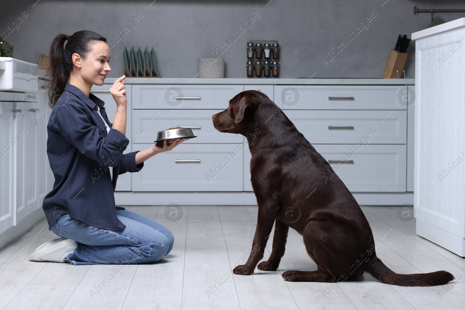 Photo of Woman with feeding bowl giving dry pet food to her dog indoors