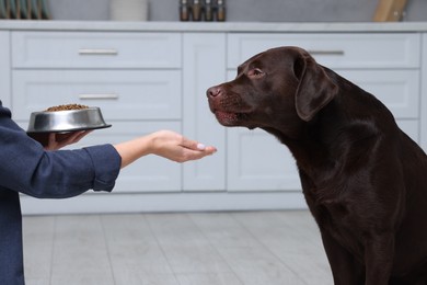 Photo of Woman with feeding bowl giving dry pet food to her dog indoors, closeup