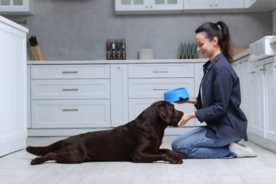 Photo of Woman with feeding bowl giving dry pet food to her dog indoors