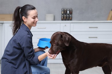 Photo of Woman with feeding bowl giving dry pet food to her dog indoors