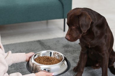 Photo of Woman giving bowl with dry pet food to her dog indoors, closeup