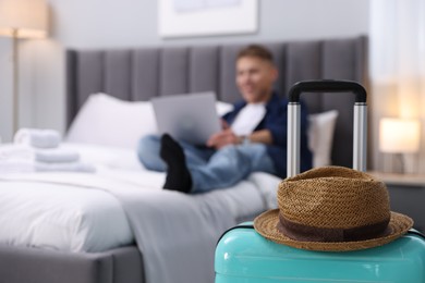 Photo of Suitcase with hat and man using laptop on bed in his hotel room, selective focus