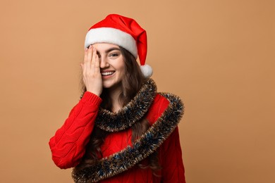 Happy young woman with tinsel and Santa hat on beige background. Space for text