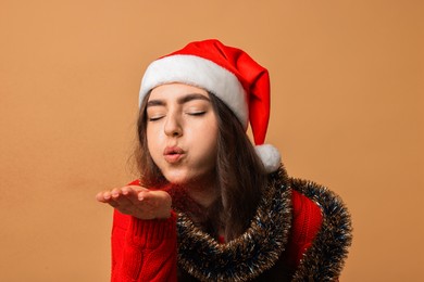 Photo of Young woman with bright tinsel and Santa hat blowing away glitters on beige background