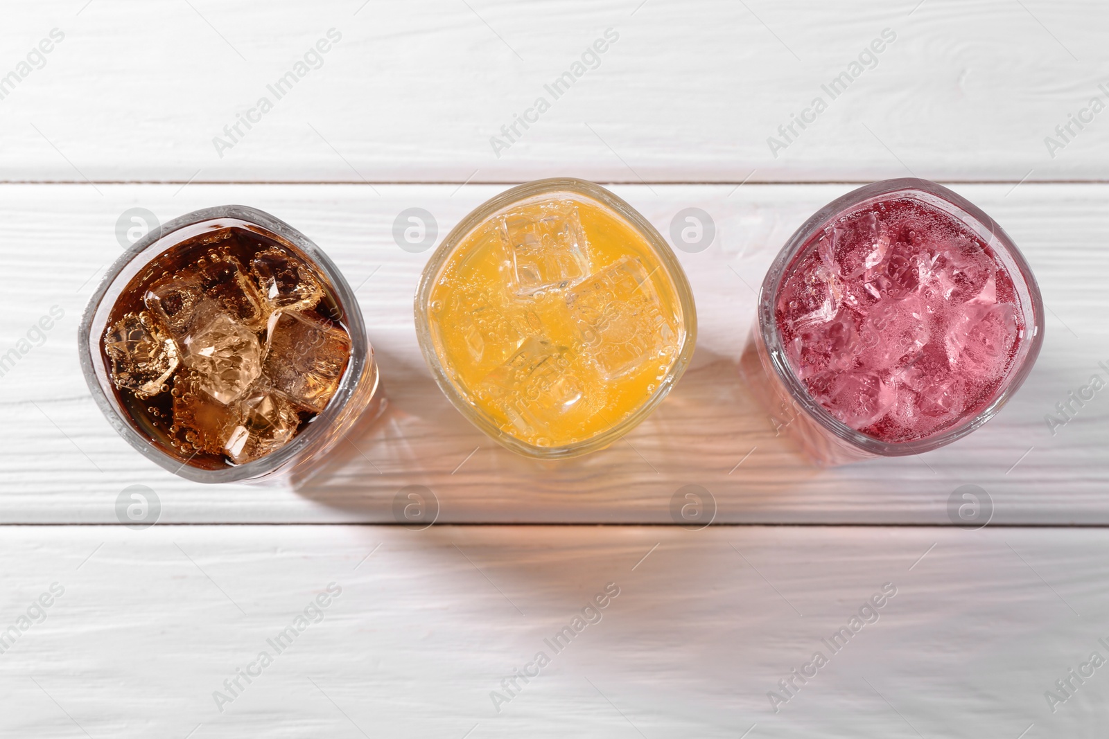 Photo of Soda water of different flavors with ice cubes in glasses on white wooden table, top view