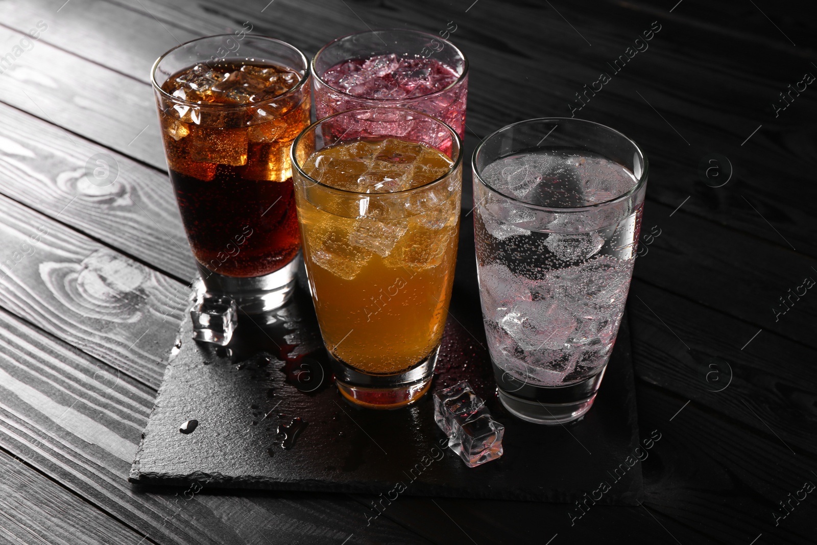 Photo of Refreshing soda water of different flavors with ice cubes in glasses on black wooden table