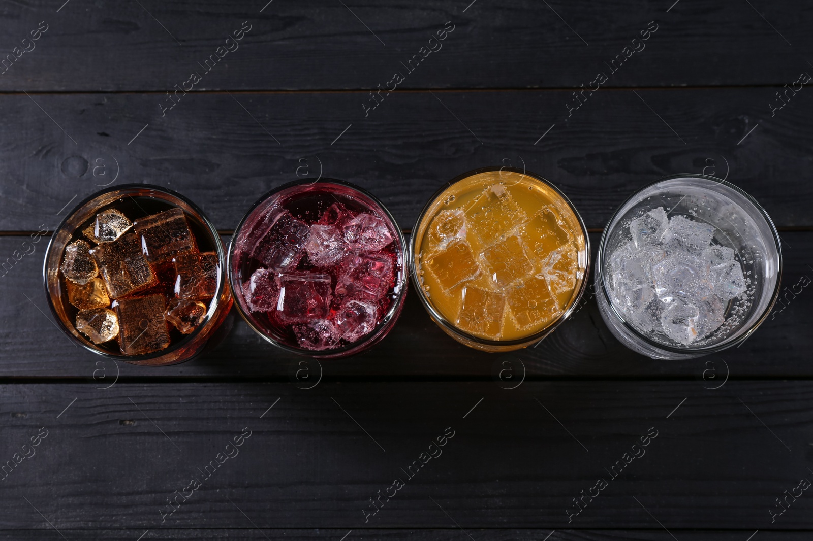 Photo of Refreshing soda water of different flavors with ice cubes in glasses on black wooden table, flat lay