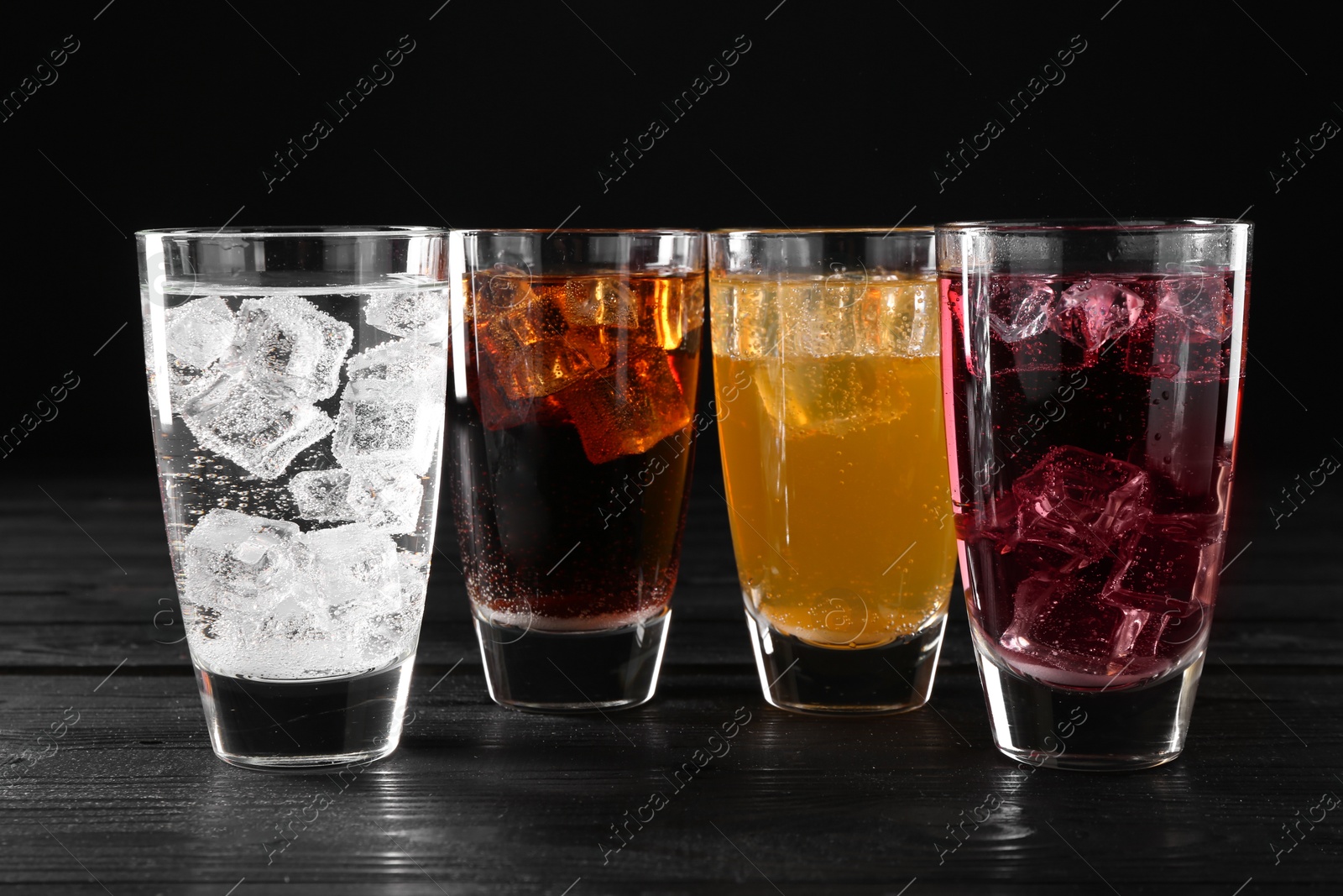 Photo of Refreshing soda water of different flavors with ice cubes in glasses on black wooden table