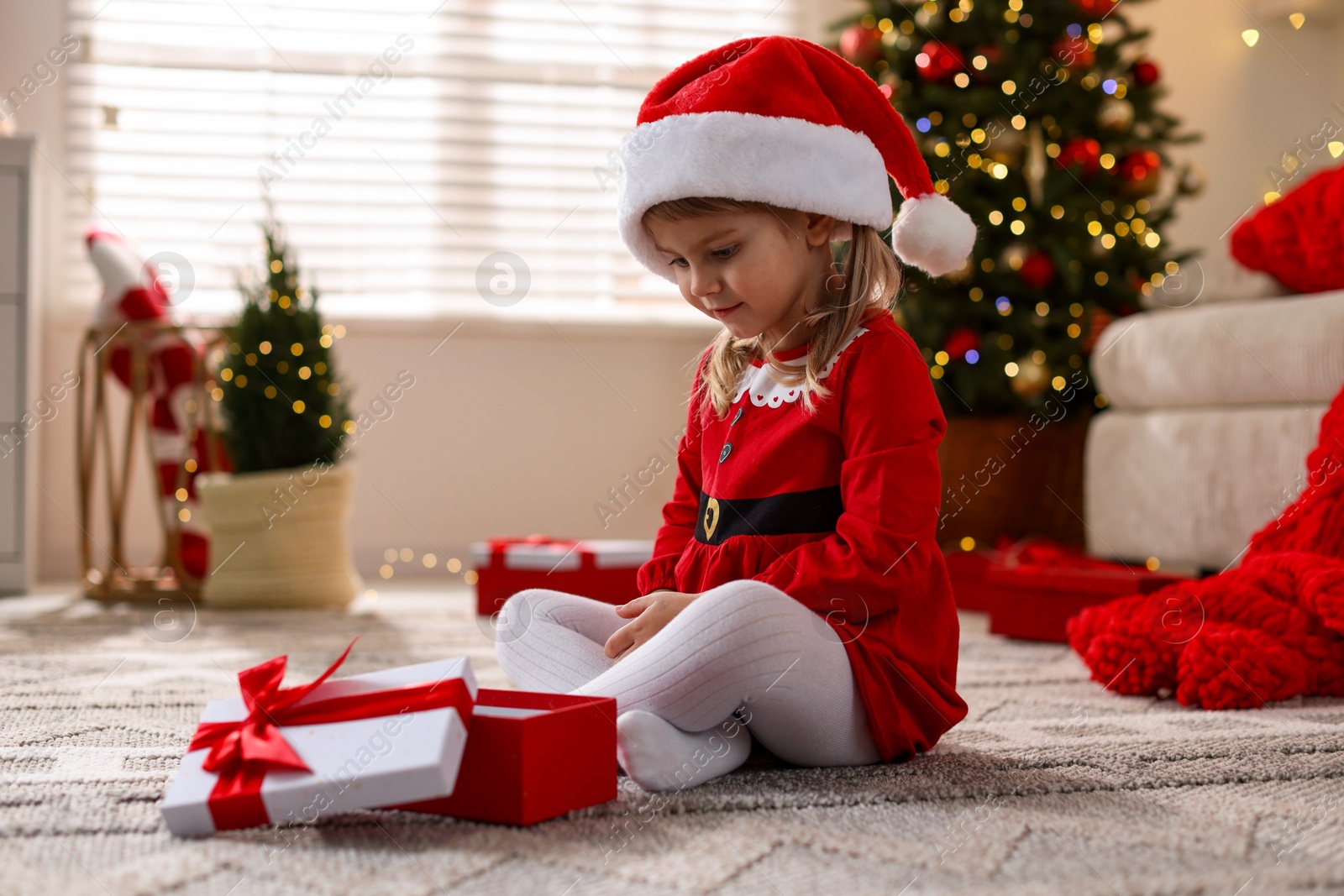 Photo of Little girl in Santa hat with gift box sitting on floor at home