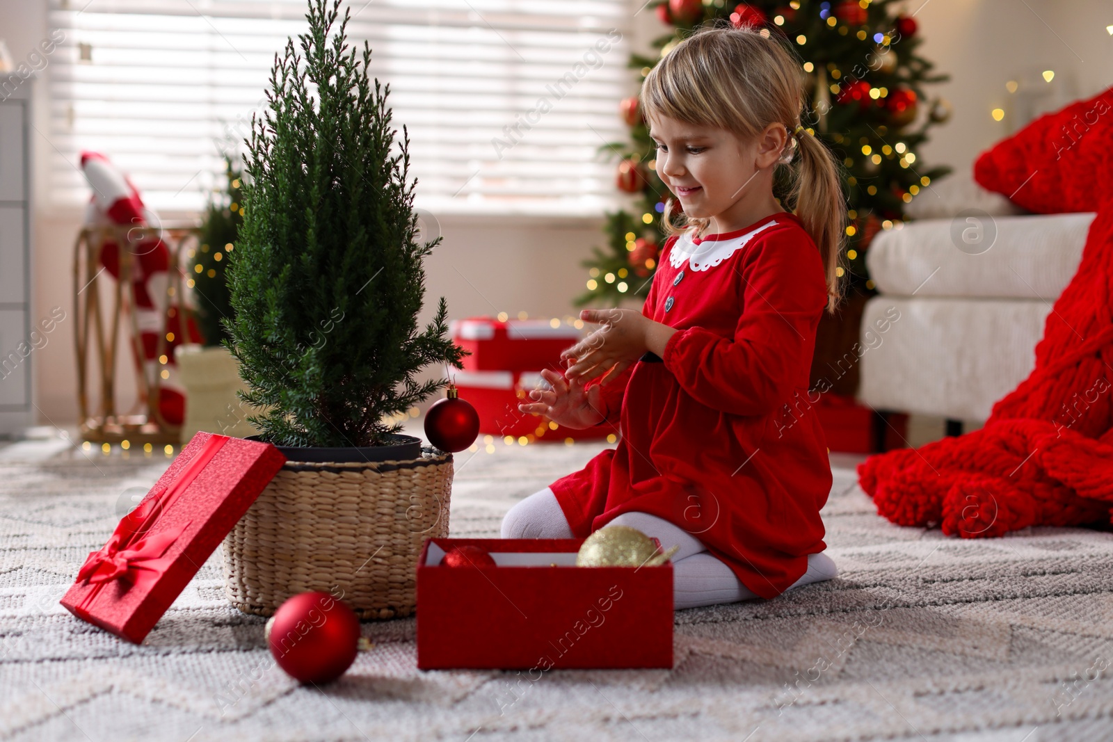 Photo of Little girl in Christmas costume with gift box decorating small tree at home