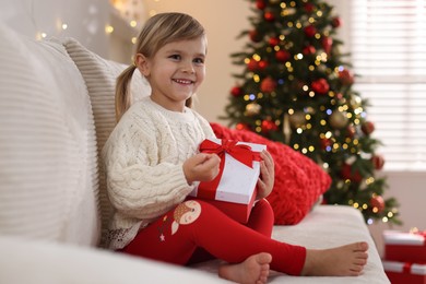 Photo of Little girl with Christmas gift box sitting on sofa at home