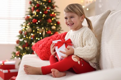 Photo of Little girl with Christmas gift box sitting on sofa at home