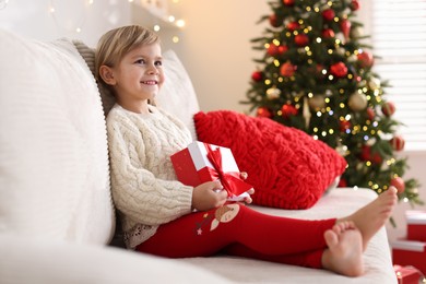 Photo of Little girl with Christmas gift box sitting on sofa at home