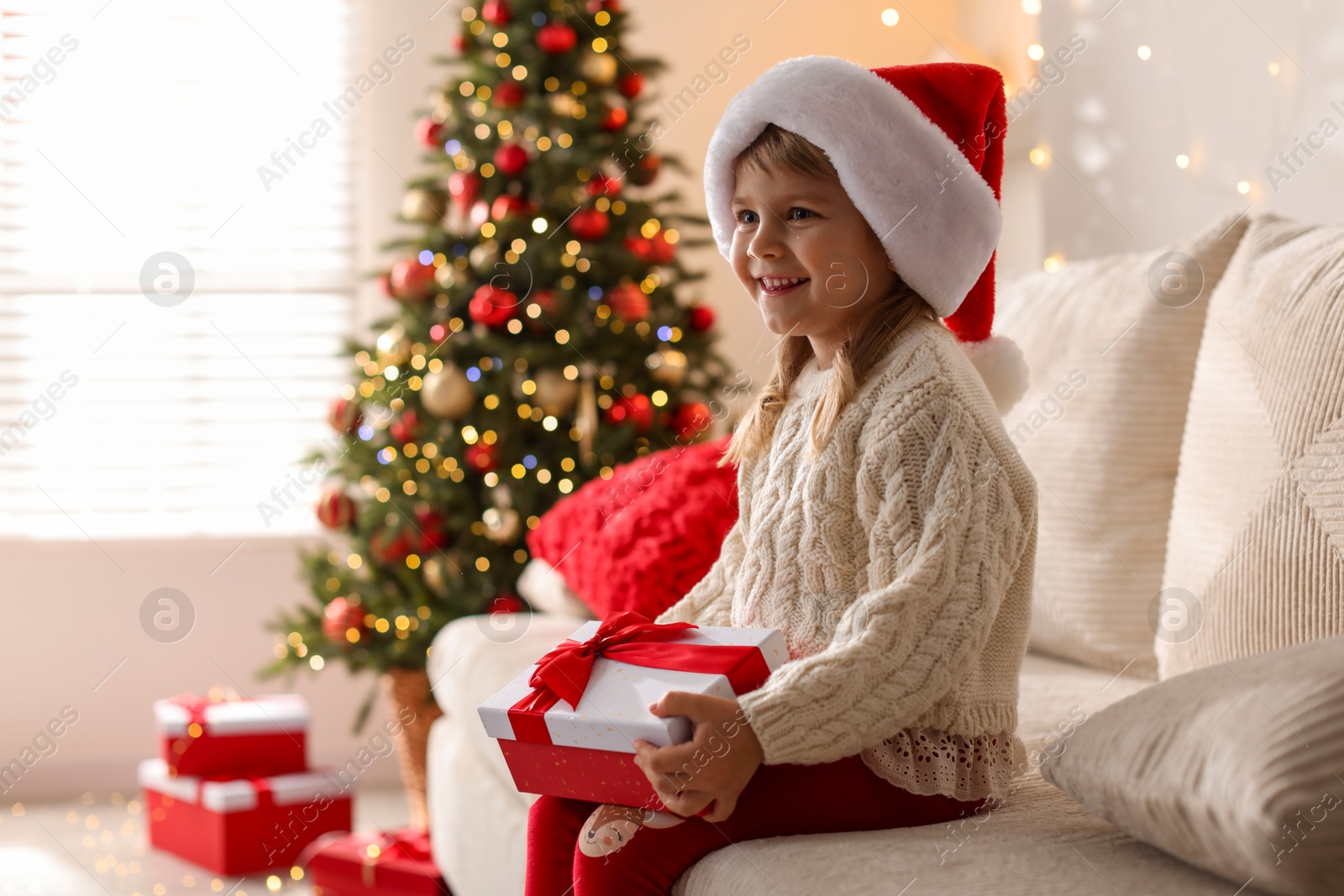 Photo of Little girl in Santa hat sitting on sofa at home