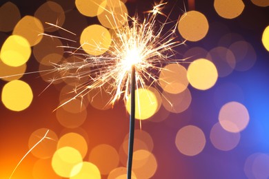 Photo of Bright burning sparkler on color background with blurred lights, bokeh effect