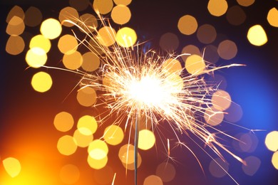 Photo of Bright burning sparkler on color background with blurred lights, bokeh effect