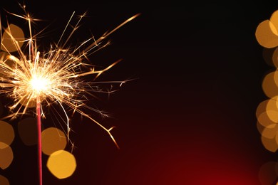 Photo of Bright burning sparkler on dark red background with blurred lights, space for text. Bokeh effect