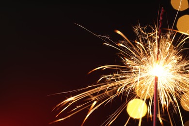 Photo of Bright burning sparkler on dark red background with blurred lights, space for text. Bokeh effect