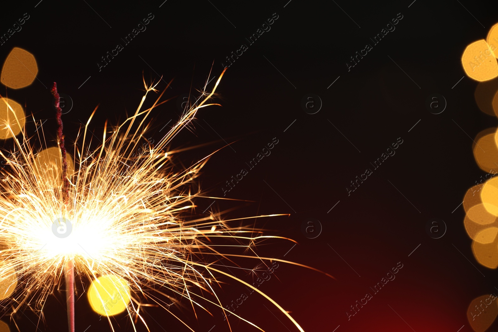 Photo of Bright burning sparkler on dark red background with blurred lights, space for text. Bokeh effect