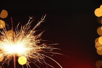 Photo of Bright burning sparkler on dark red background with blurred lights, space for text. Bokeh effect