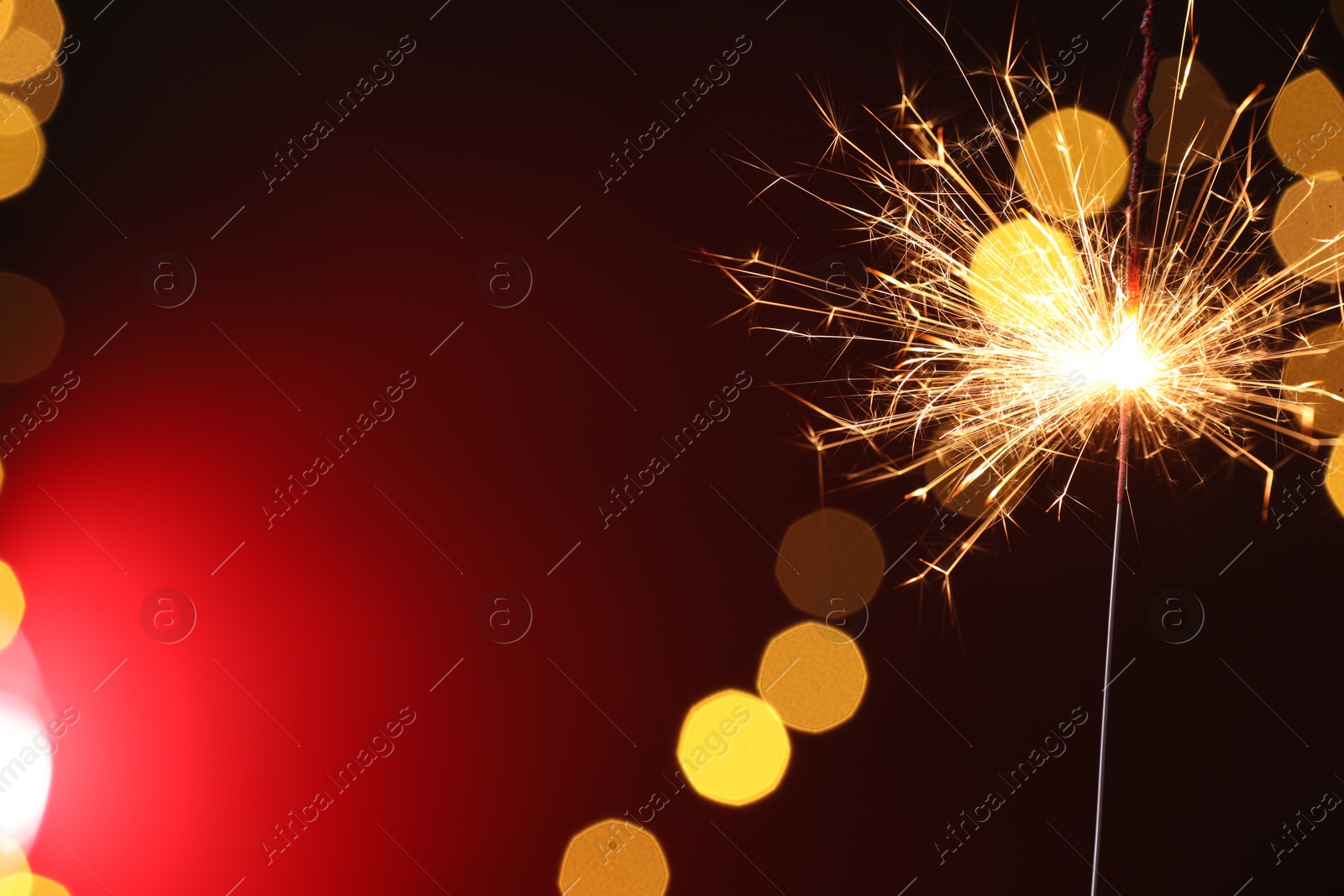 Photo of Bright burning sparkler on dark red background with blurred lights, space for text. Bokeh effect