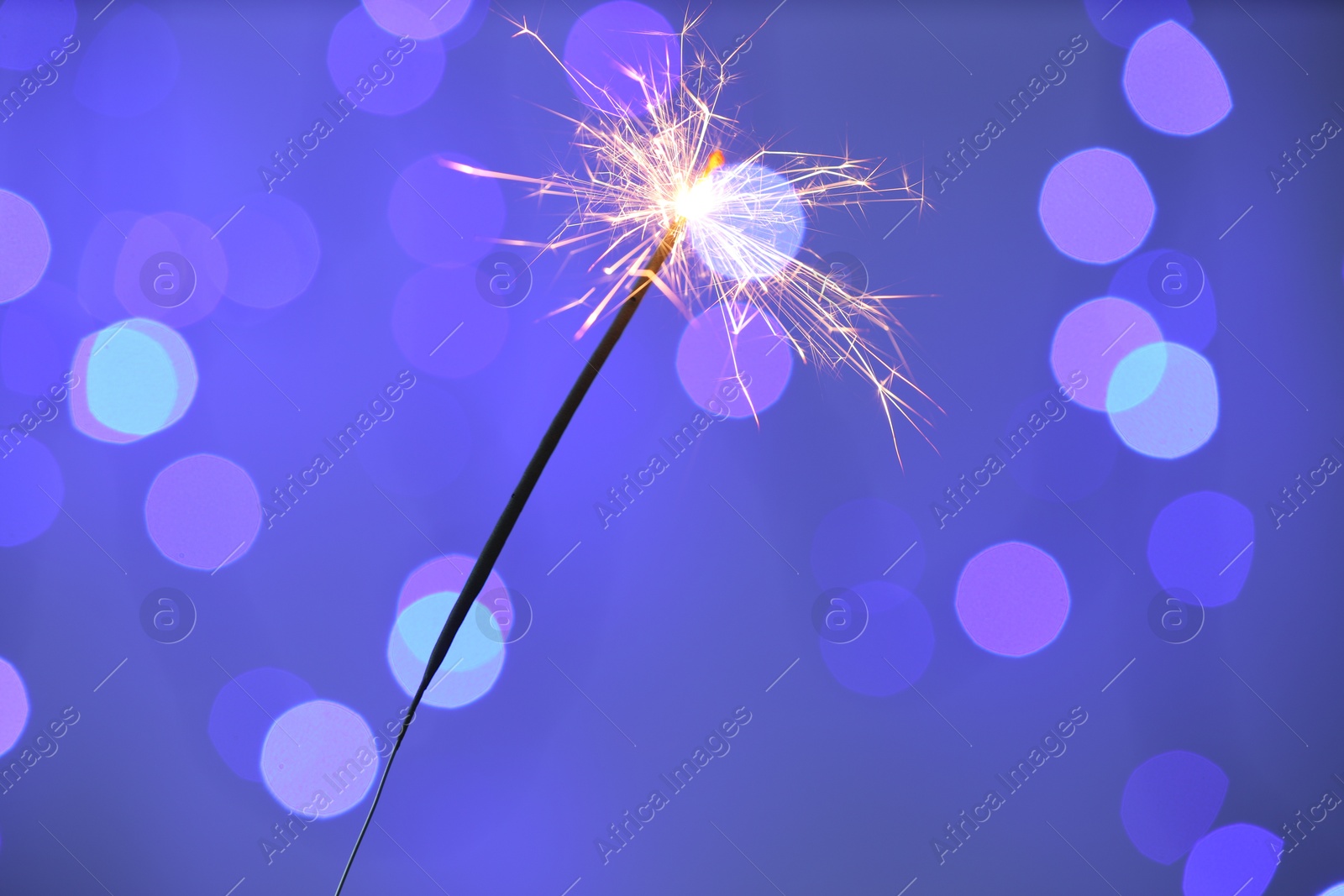 Photo of Bright burning sparkler on color background with blurred lights, bokeh effect