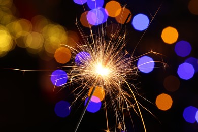 Photo of Bright burning sparkler on dark background with blurred lights, bokeh effect