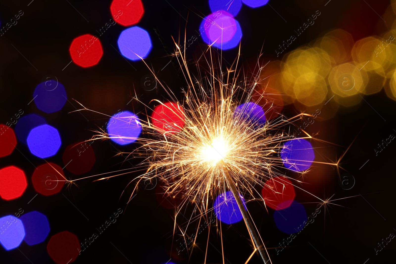 Photo of Bright burning sparkler on dark background with blurred lights, bokeh effect