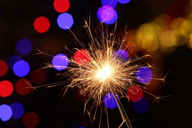 Photo of Bright burning sparkler on dark background with blurred lights, bokeh effect