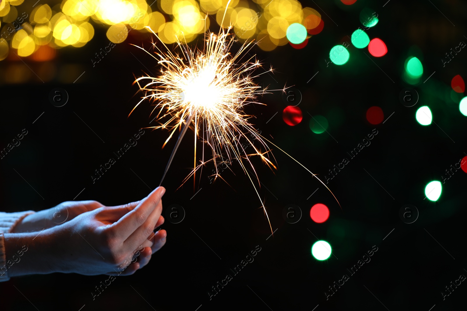 Photo of Man holding bright burning sparkler on dark background with blurred lights, closeup. Bokeh effect