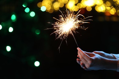 Photo of Man holding bright burning sparkler on dark background with blurred lights, closeup. Bokeh effect