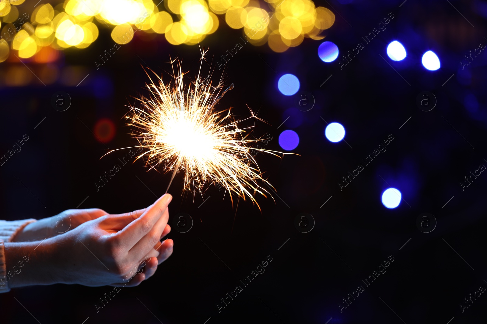 Photo of Man holding bright burning sparkler on dark background with blurred lights, closeup. Bokeh effect