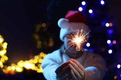 Photo of Man holding bright burning sparkler on dark background with blurred lights, selective focus. Space for text