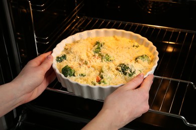 Photo of Woman putting uncooked vegetable casserole into oven, closeup