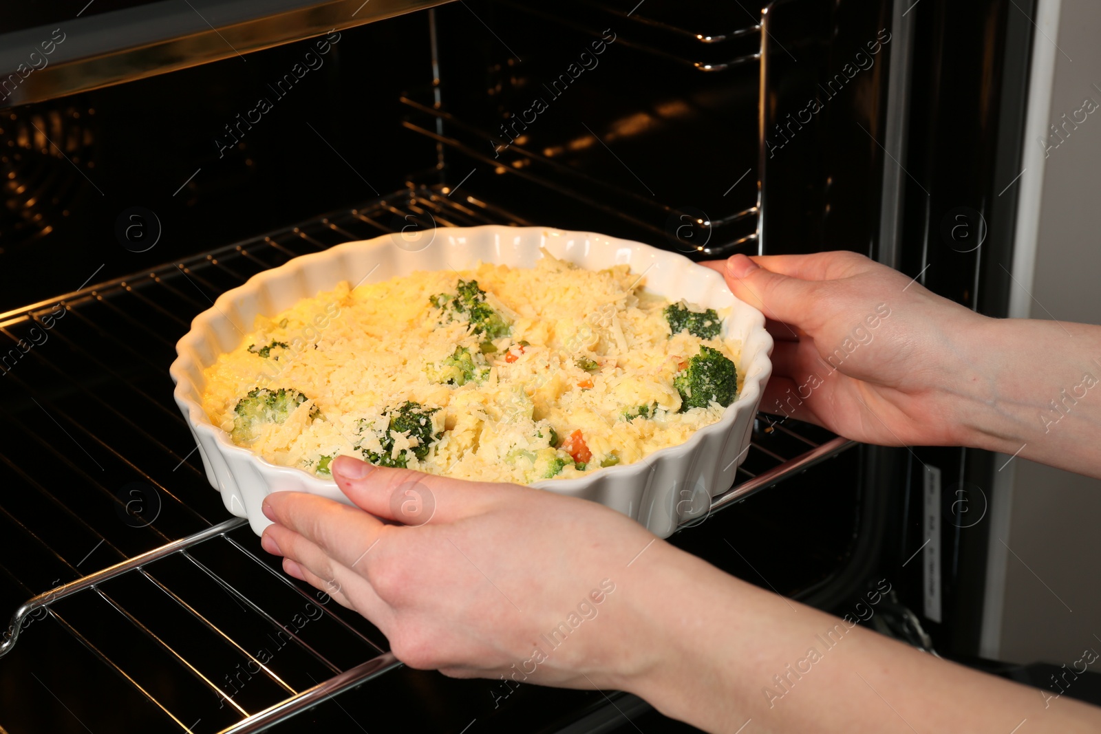 Photo of Woman putting uncooked vegetable casserole into oven, closeup