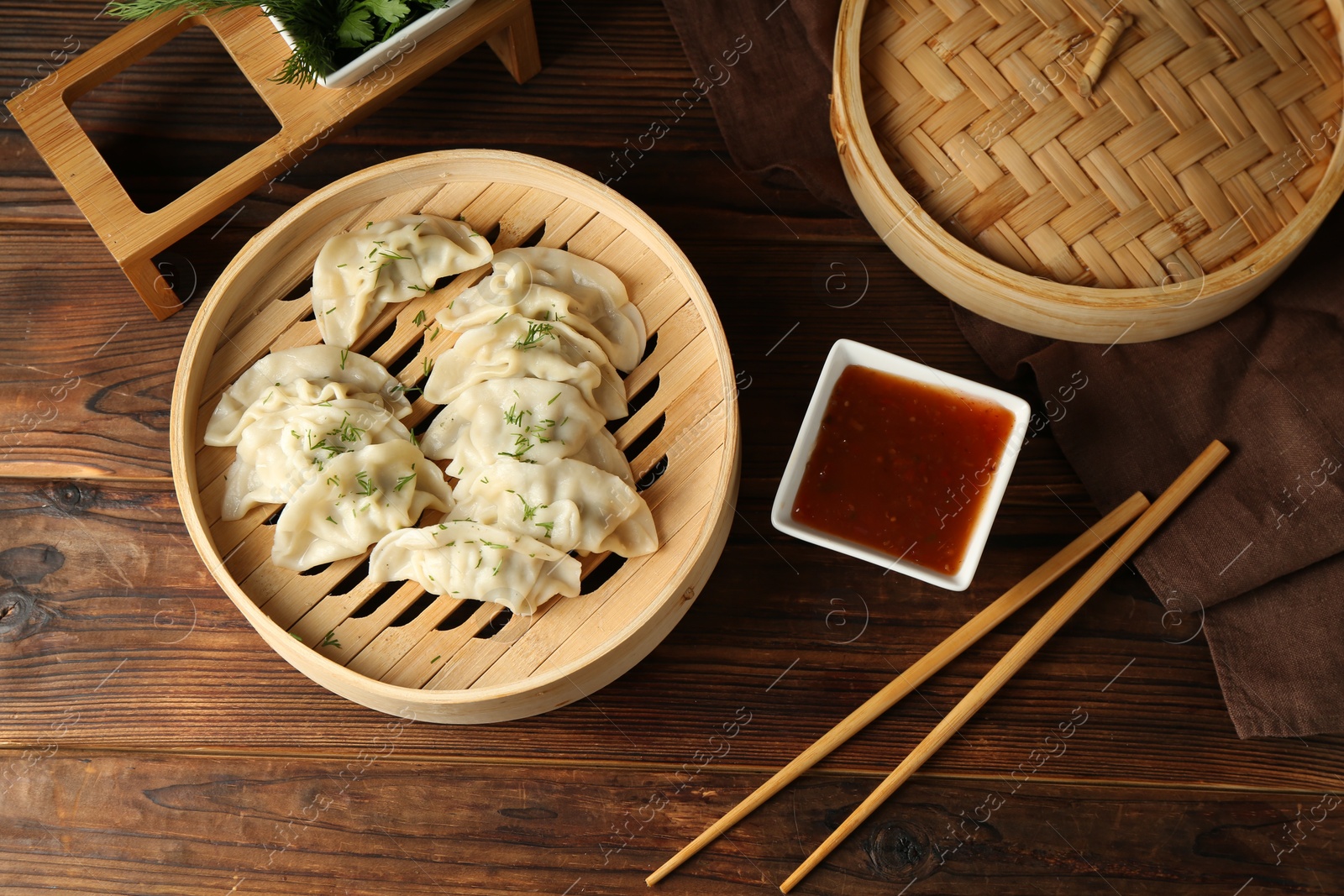 Photo of Tasty boiled gyoza (dumplings) in bamboo steamer, sauce and chopsticks on wooden table, flat lay