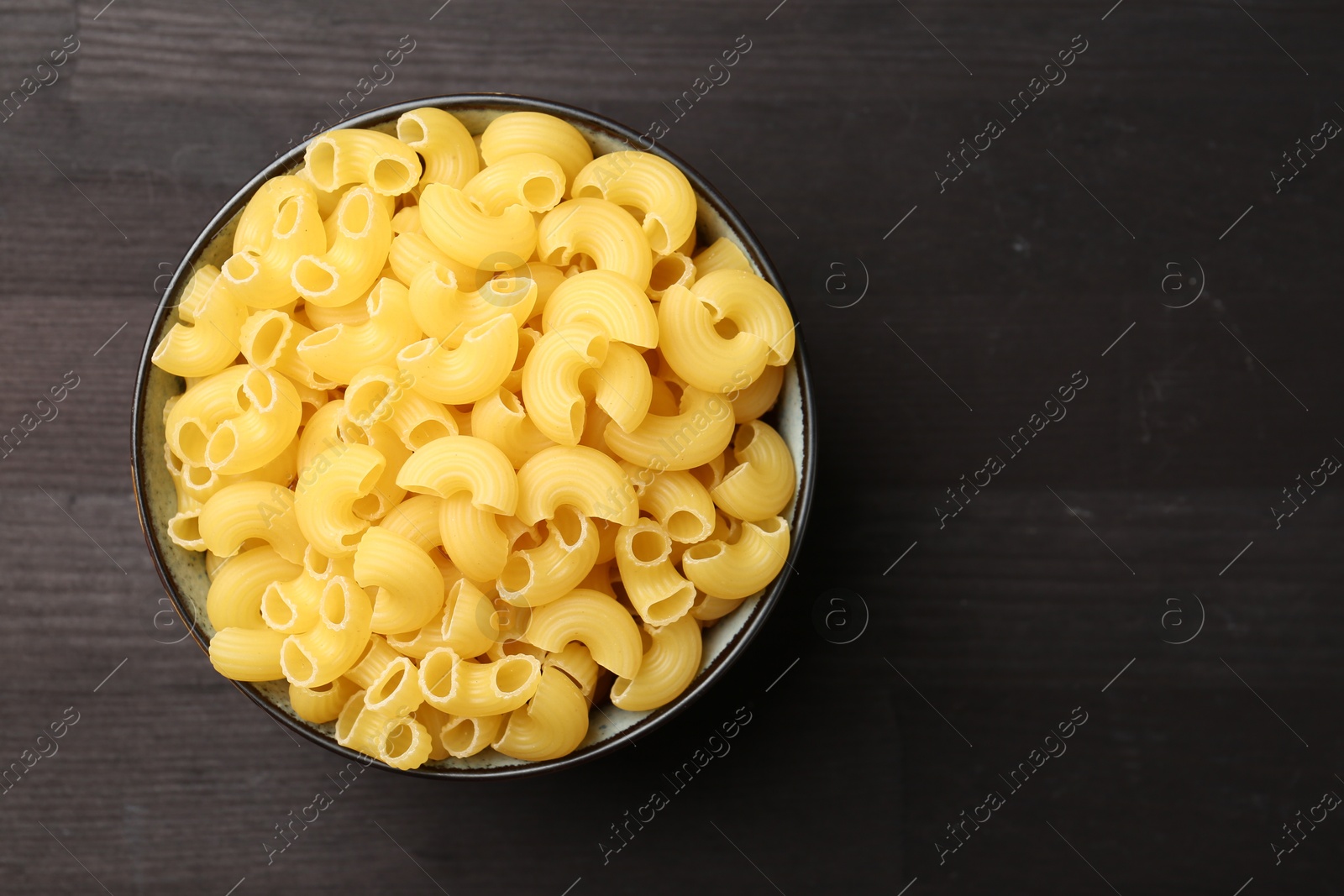 Photo of Raw horns pasta in bowl on dark wooden table, top view. Space for text