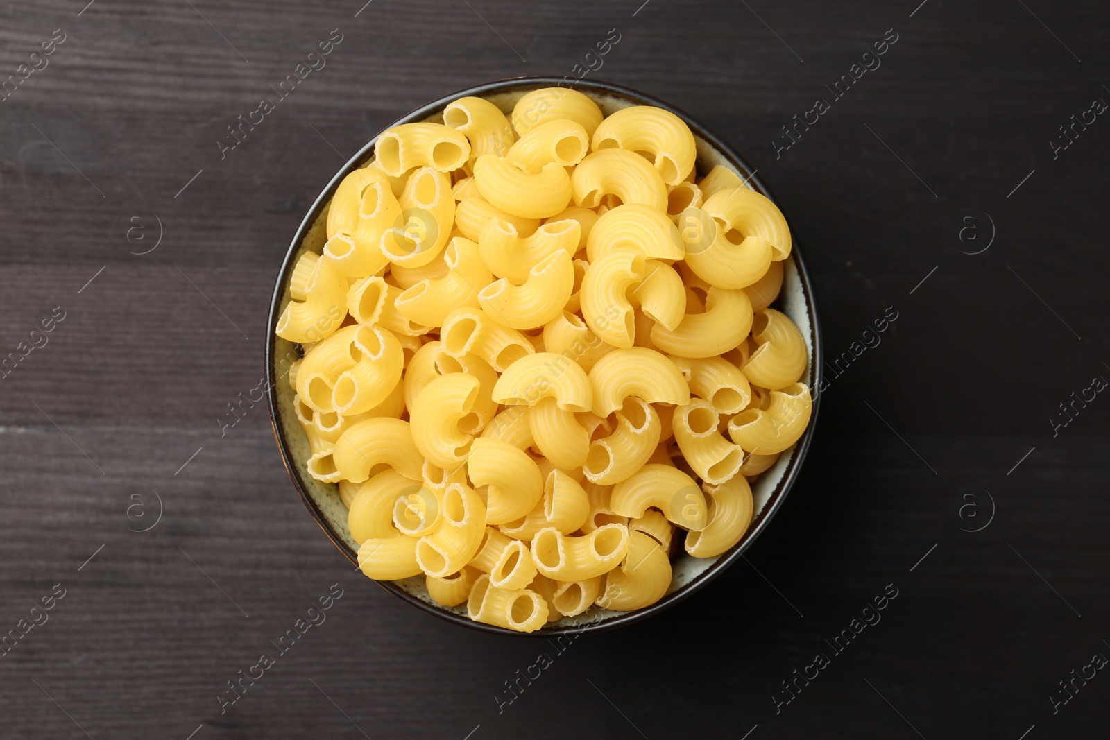 Photo of Raw horns pasta in bowl on dark wooden table, top view