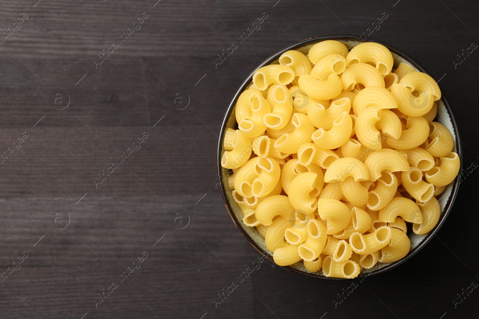 Photo of Raw horns pasta in bowl on dark wooden table, top view. Space for text