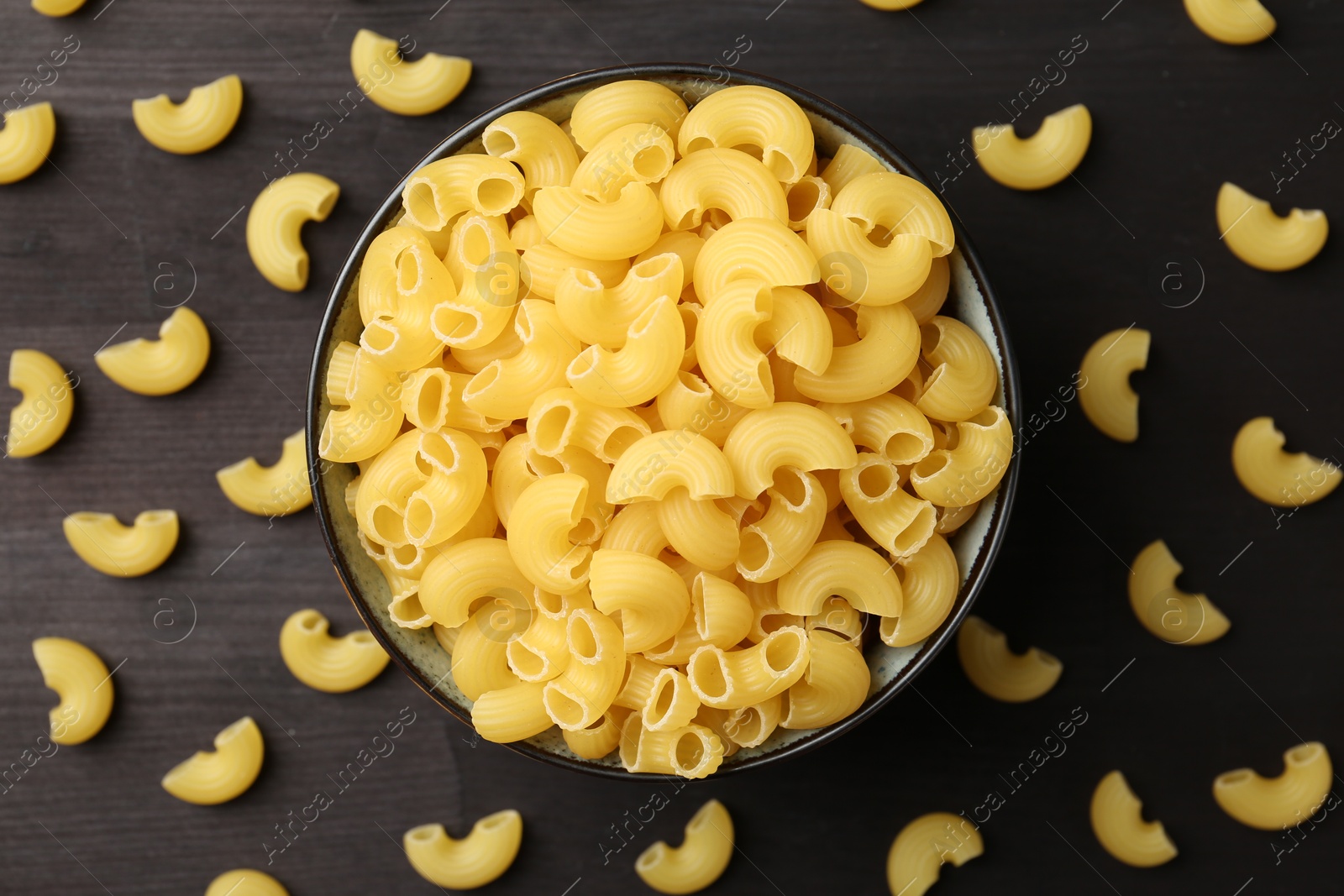 Photo of Raw horns pasta in bowl on dark wooden table, top view