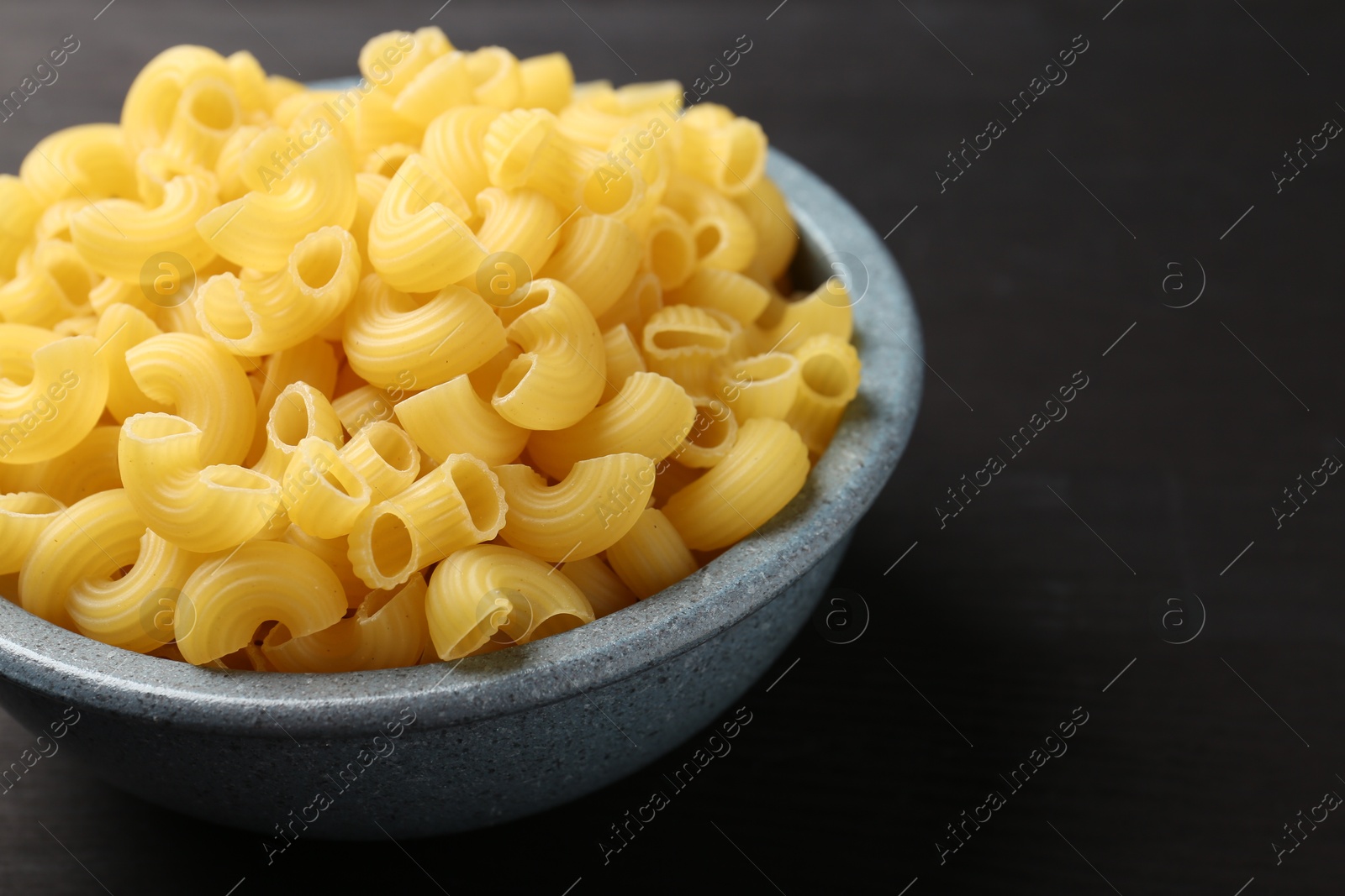 Photo of Raw horns pasta in bowl on dark table, closeup