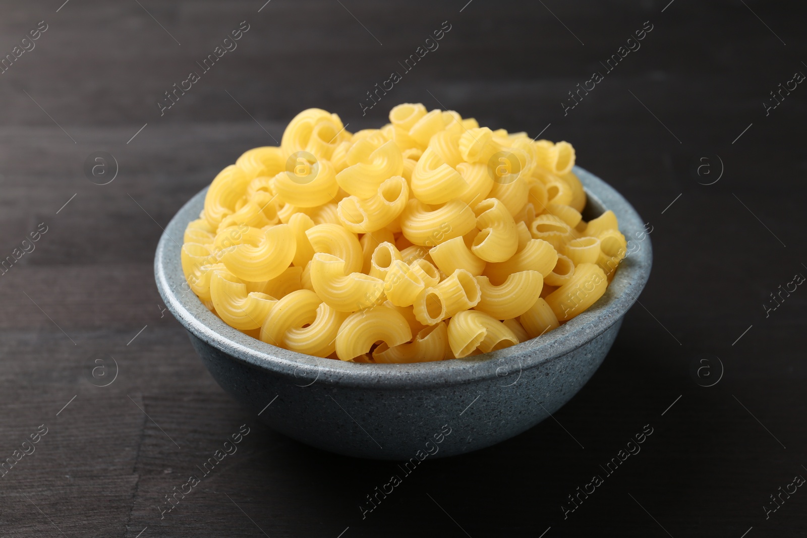 Photo of Raw horns pasta in bowl on dark wooden table, closeup