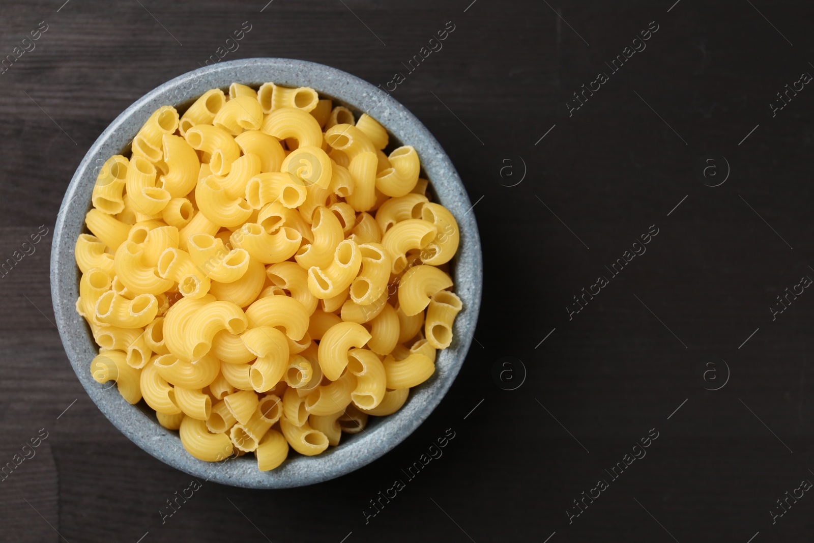 Photo of Raw horns pasta in bowl on dark wooden table, top view. Space for text