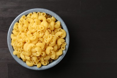 Photo of Raw horns pasta in bowl on dark wooden table, top view. Space for text