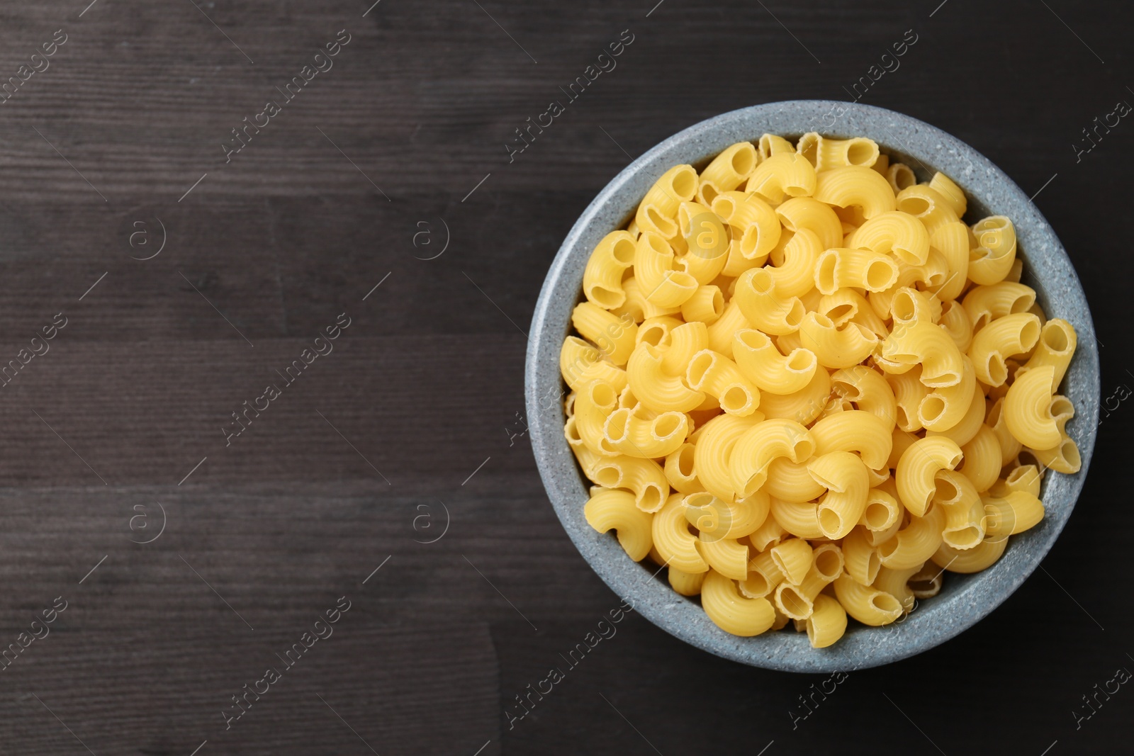 Photo of Raw horns pasta in bowl on dark wooden table, top view. Space for text