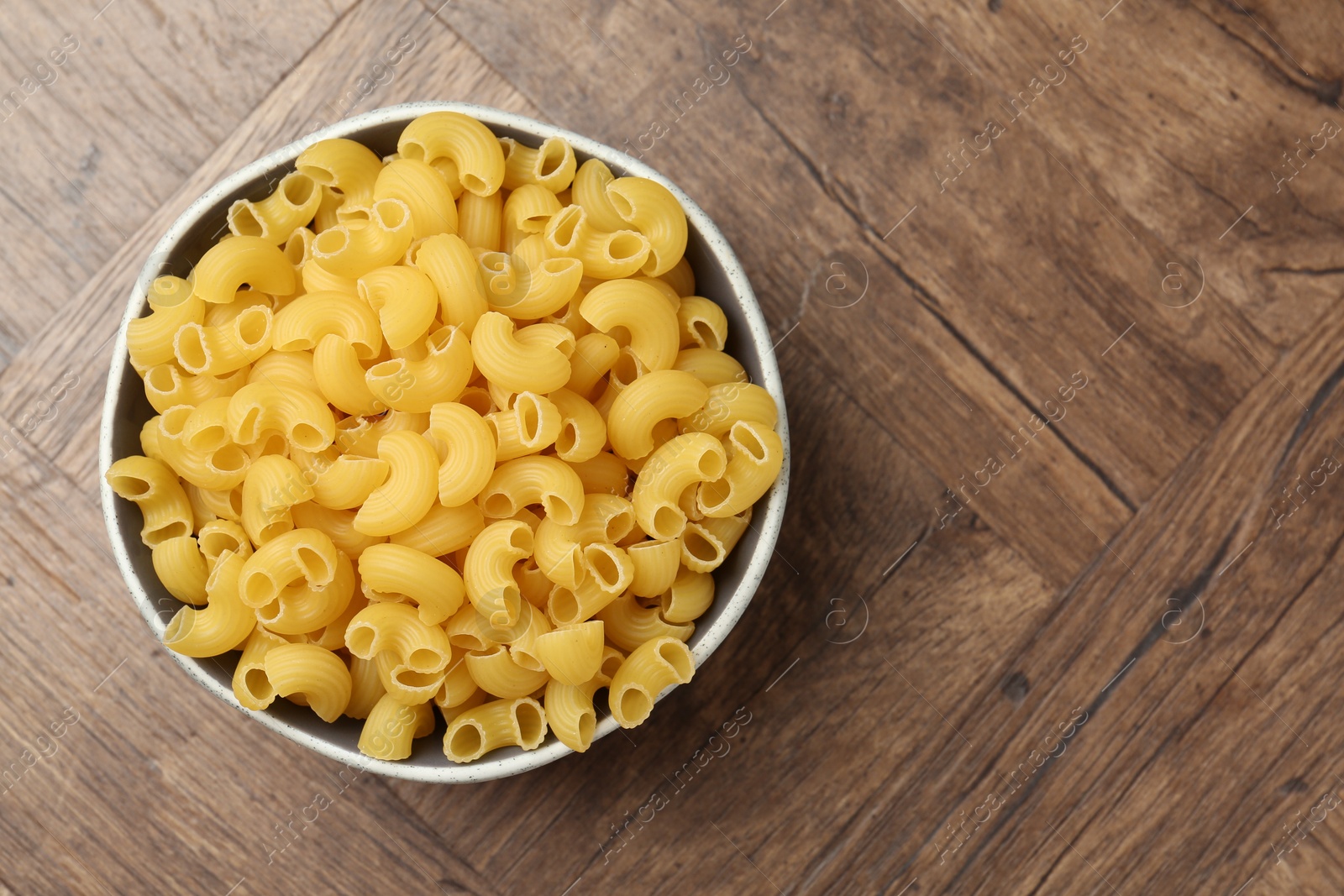 Photo of Raw horns pasta in bowl on wooden table, top view. Space for text