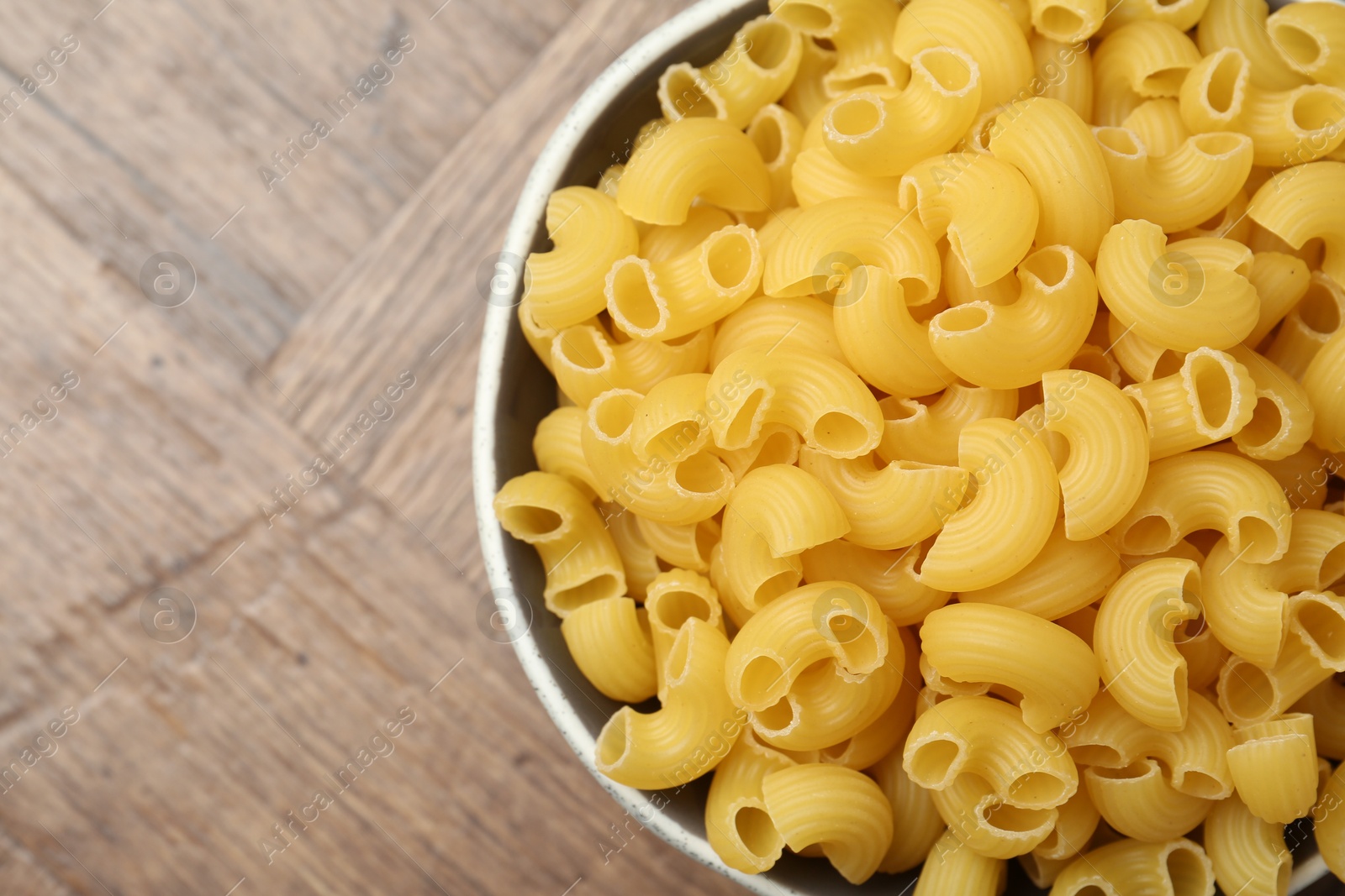 Photo of Raw horns pasta in bowl on wooden table, top view. Space for text